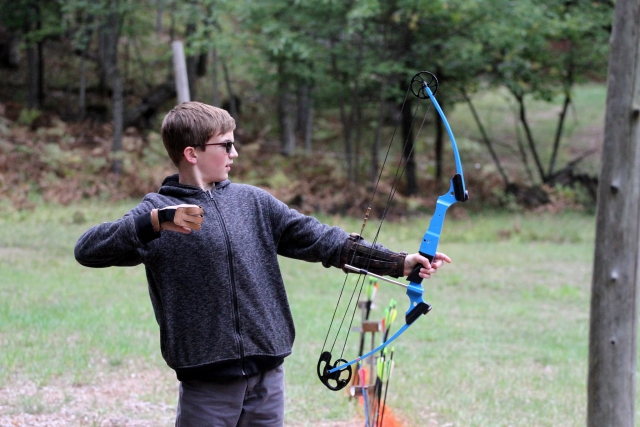Young boy wearing sunglasses and grey sweater doing archery