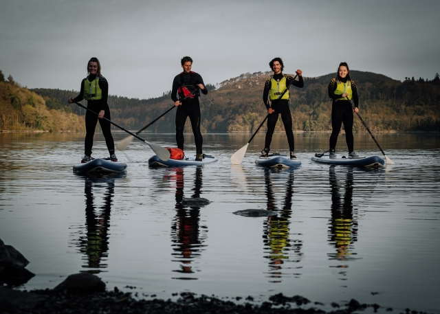  Quattro persone che indossano mute nere, aiuti al galleggiamento verdi paddleboarding al lago 