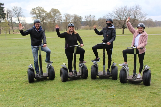 Four friends weatring blak helmets raising their hand and smiling while doing segway in the park