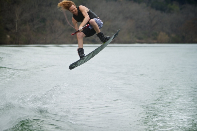 garoto vestindo auxílios pretos de flutuabilidade pulando uma onda enquanto faz wakeboard 