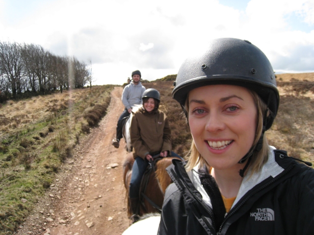 Lächelndes Mädchen mit schwarzem Helm und Pullover und zwei Freunden im Rücken beim Reiten 