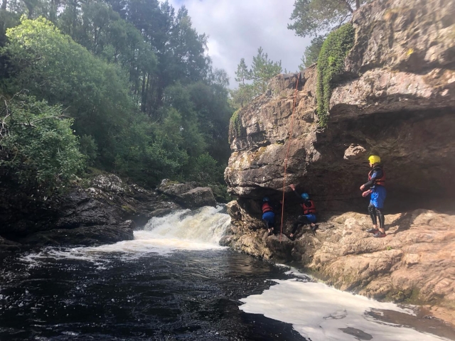Three people wearing yellow helmets and red buoyancy aids caving and climbing during their canyoning session