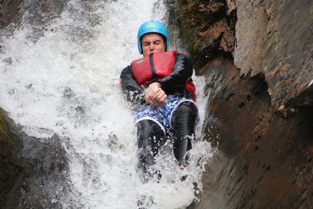  Junge schließt die Augen, trägt blauen Helm und rote Schwimmhilfen und springt von den Wasserfällen 