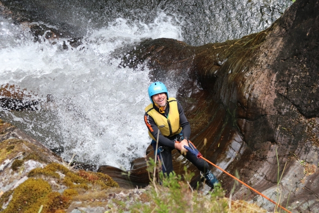  Uomo sorridente e vestito Casco blu e salvagente giallo durante l'arrampicata durante la sua sessione di canyoning 