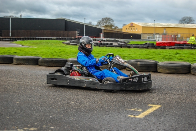 Jeune garçon portant une combinaison de conduite bleue et un casque noir de karting avec des pneus le long de la piste 