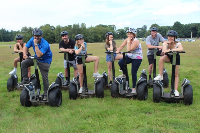 Group of boys and girl wearing t-shirts, grey helmets, smiling and doing segway around the park