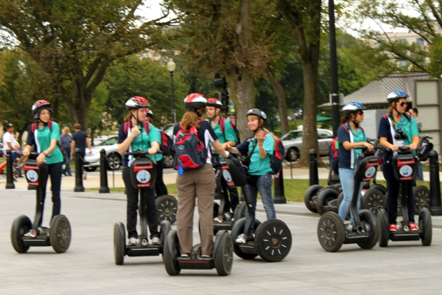 People wearing blue shirts and red and white helmets riding segways in the park